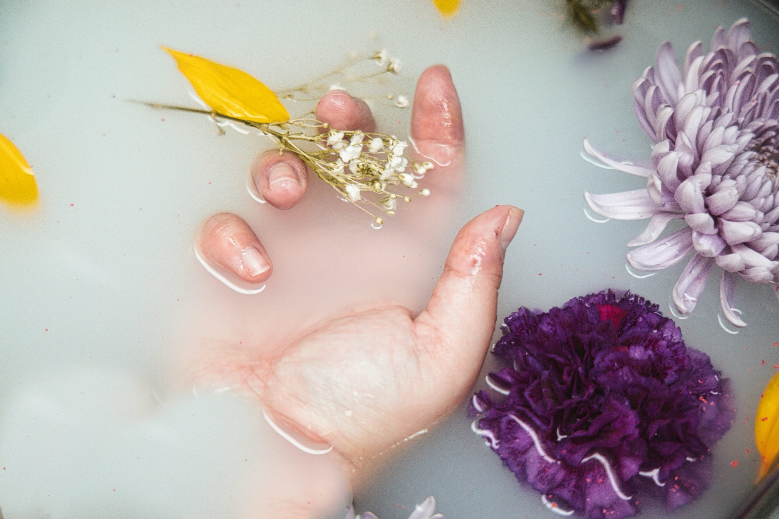 Đôi bàn tay đang được ngâm trong bát nước ấm với cánh hoa, giúp làm mềm da và thư giãn móng tay.